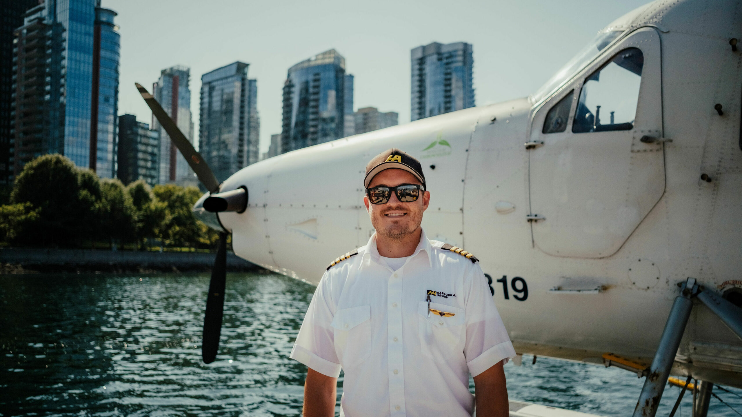 A pilot smiles in front of his plane, parked in front of a scenic city harbour.