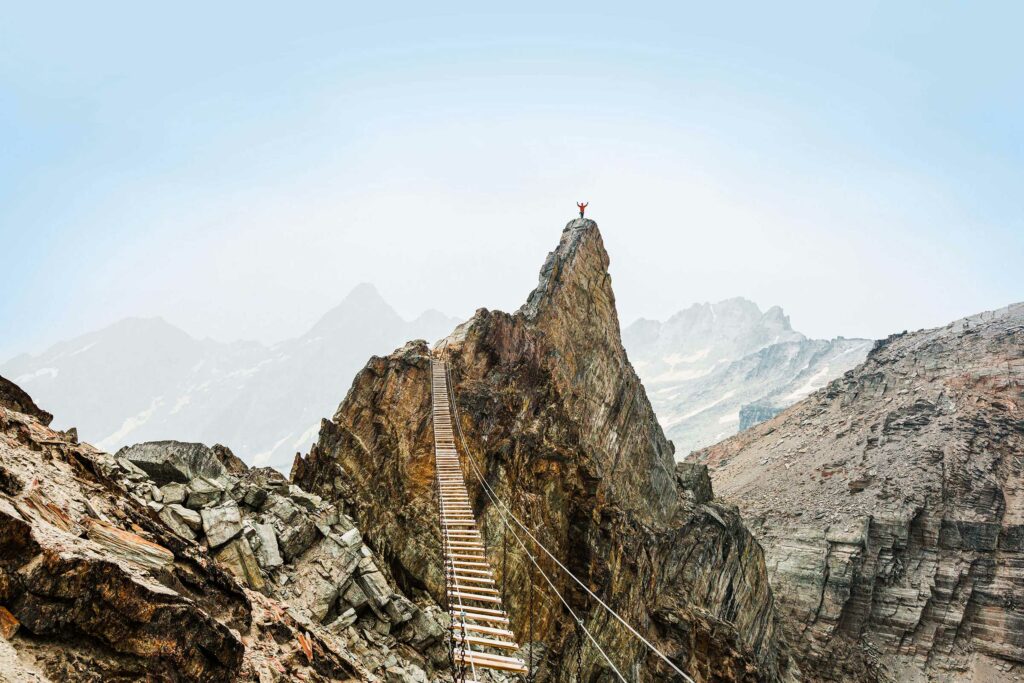 Un pont suspendu en bois relie des pics et mène à une personne qui se tient, triomphante, au sommet de la montagne.