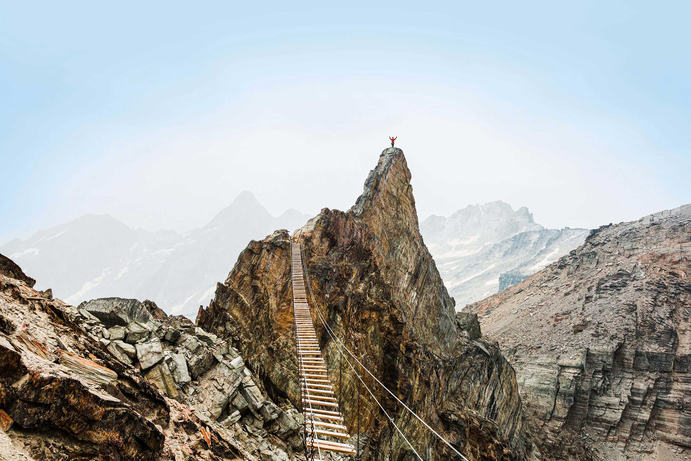 Wooden suspension bridge spanning across mountain peaks leading to a person triumphantly standing atop the mountain.