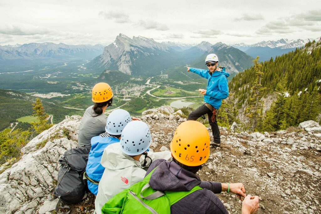 Person talking to a group of four on a moutain. Person is pointing at another moutain while the group of four are listening to him. 
