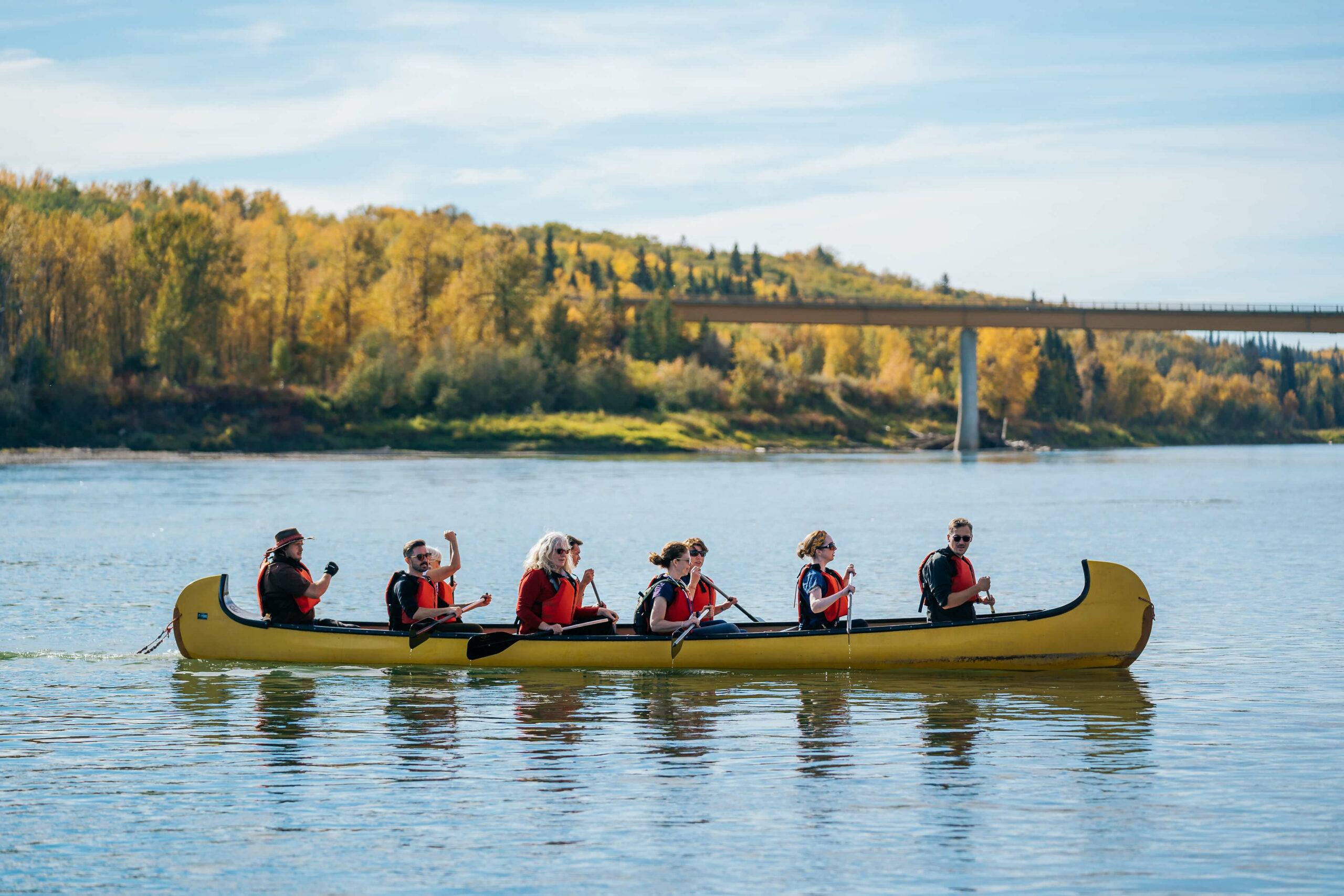 Nine people in a canoe on a calm lake with a forest and bridge in the background.