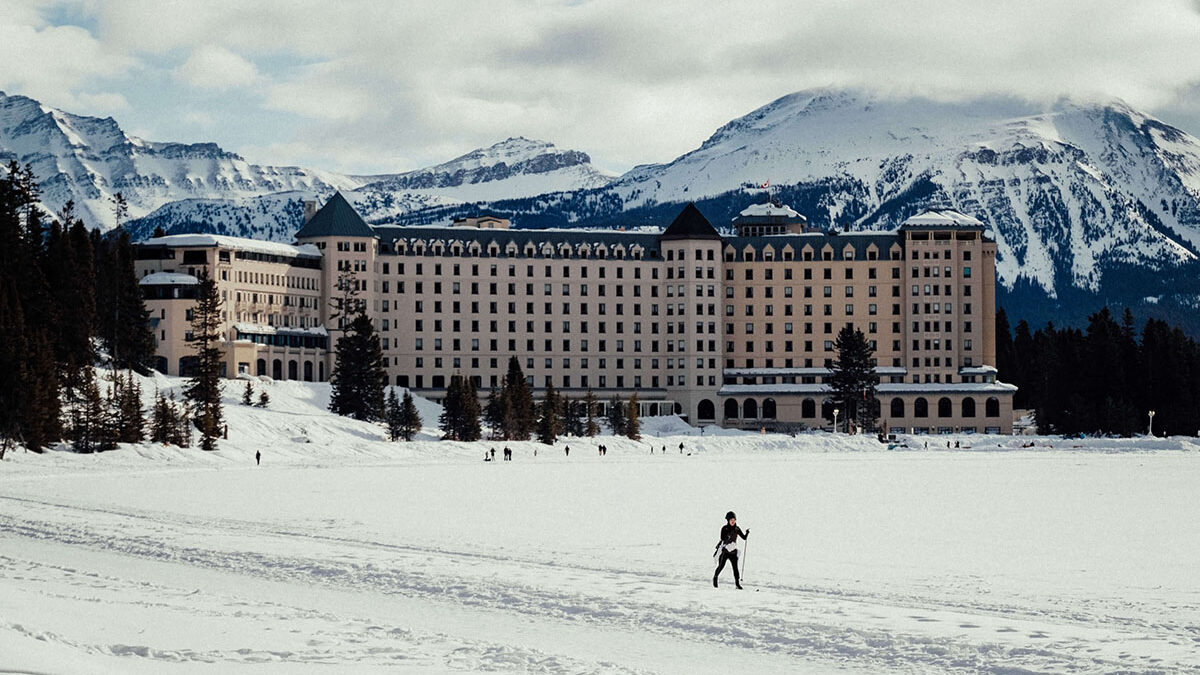 Person cross country skiing in the middle distance with a large hotel and mountain range in the background.