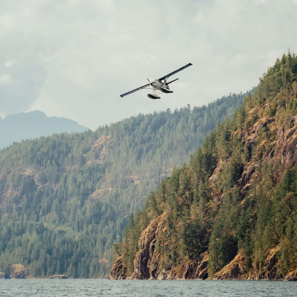 Seaplane flying above a lake surrounded by forested mountains.