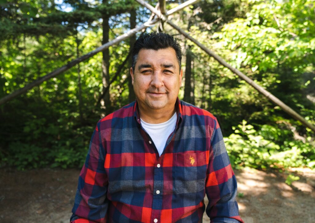 Person standing in the forest beneath wood shelter frame.